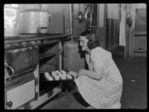 Portrait of an unidentified female TEAL Catering Staff member putting a tray of scones into an oven within the kitchen of an unidentified building, [Auckland?]