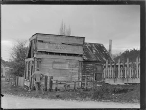 Goldmining, view of the remains of an old single story wooden bank building with an unidentified man in front, South Westland, West Coast Region