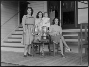 Bristol Freighter tour, Hawkes Bay and East Aero Club, showing Miss J Penny, Mrs Temple Martin (I/C Clubhouse), small child and Miss N Dunn, standing on a house porch, with trophy cups won at the New Zealand Pageant at New Plymouth