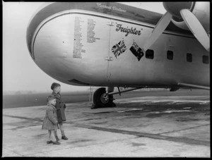 The two young sons of Leo White, under the nose of a Bristol Freighter 'Merchant Venturer' aircraft, including a list of airports visited on the aircraft and a British and New Zealand flag, location unidentified