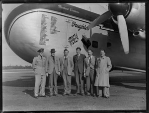 Group portrait of crew members of the visiting Bristol Freighter transport plane 'Merchant Venturer' G-AIMC with names of airports visited on front cargo door, Whenuapai Airfield, Auckland