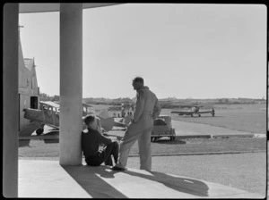 Two unidentified men on the steps of the clubrooms at Auckland Aero Club, Mangere, Manukau City, showing a view of the airfield and hangar