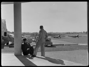 Two unidentified men on the steps of the clubrooms at Auckland Aero Club, Mangere, Manukau City, Auckland Region, including a view of aircraft and airfield