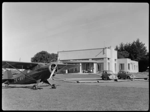 View of the clubrooms at Auckland Aero Club, Mangere, Auckland, showing Rearwin Sportster aeroplane ZK-AKF and two motorcars parked outside