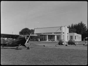 Scene at Auckland Aero Club, Mangere, Manukau City, Auckland Region, including unidentified people sitting on steps of clubrooms, a Rearwin Sportster aeroplane ZK-AKF, and parked motorcars