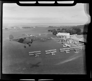 Rearwin Sportster aeroplane ZK-AKF, in flight over Auckland Aero Club, Mangere, Manukau City, Auckland Region, showing clubrooms, hangar, and a group of de Havilland Tiger Moth aeroplane lined up on ground