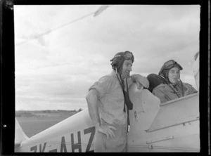 Mr K Robinson and Miss Kelsey, both wearing aviator helmets, with a de Havilland Tiger Moth aeroplane ZK-AHZ, probably at Auckland Aero Club, Mangere, Auckland