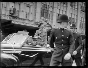 Lord Montgomery standing in car waving to people in street parade, with policemen alongside vehicle, [Queen Street?], Auckland