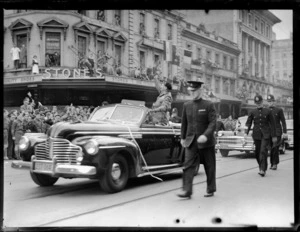 Lord Montgomery standing in car waving to people in street parade, with policemen alongside vehicle, outside Stone's shoe shop, [Queen Street?], Auckland