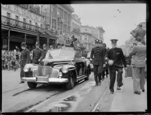 Lord Montgomery standing in car, waving to people in street parade, with policemen marching alongside vehicle, outside The Strand Theatre, Queen Street, Auckland