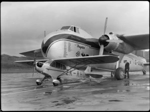 Bristol Freighter Tour, view of an Auster J/1 Autocrat ZK-APO plane in front of Bristol Freighter transport plane 'Merchant Venturer' G-AIMC, with an unidentified man looking at a Bristol Freighter wheel, Rongotai Airport, Wellington City