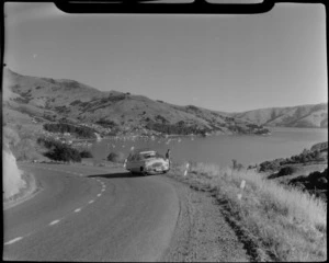 Akaroa, Banks Peninsula, showing unidentified young man with his motorcar on a road, including houses and the harbour in the distance