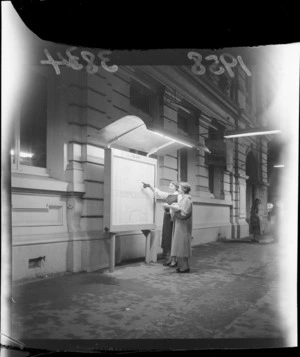 Evening scene featuring unidentified women at illuminated information shelter containing Palmerston North City guides, Manawatu-Whanganui Region