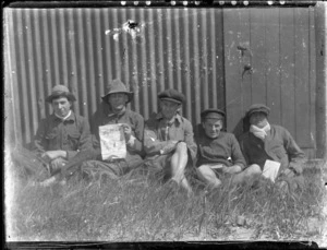 Group of men in front of corrugated iron wall - taken by an unknown photographer