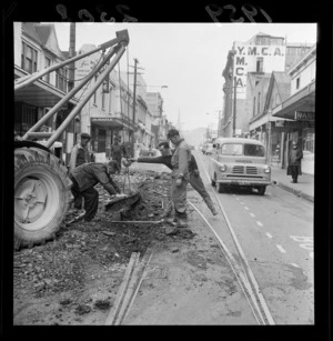 Roadworks on Willis Street, showing unidentified men moving a tram track with a crane, and view of business premises including Maple Furnishing Company Ltd and YMCA building