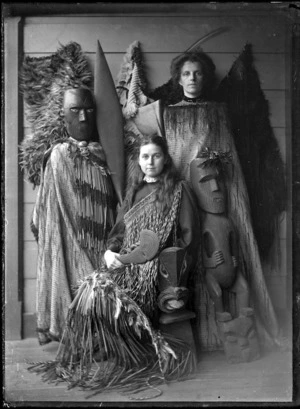 Two unidentified European women in a display of Maori cloaks, weapons, implements, and wooden carved figures.