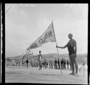 Members of Surf Life Saving Clubs on the beach at Titahi Bay during championships with banners indentifying clubs, Wellington
