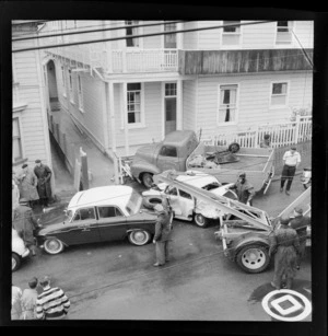 Truck and car accident, The Terrace, Wellington, showing truck smashed into a fence of a two storey building, including a tow truck, taxi cab and people standing around