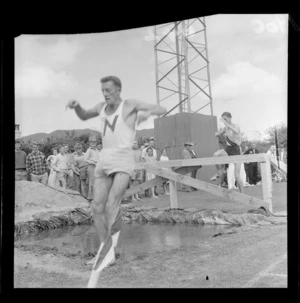 Athletics at Hataitai Park with unidentified male running competitor at a steeplechase water jump with a crowd looking on, Wellington City