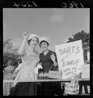 Unidentified Miss Upper Hutt contestant and an unidentified woman, at a garden party, probably Wellington, including a sign reading 'Darts, 3 Shots, 2 Scores Will Win A Prize'