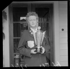 Mrs C Setter, champion bowler, with a trophy and badges on her blazer, including a patch which says 'NZWBA, National Champion of Champions, Singles 1960'