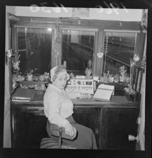 Mrs M Bowden in the Wellington Railway Station announcing box, with small pots of flowers around the office