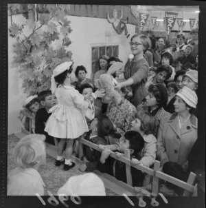 Children's fashions at Farmers Department Store with an unidentified woman with a microphone giving a young girl model a teddy bear with people looking on, probably Wellington Region