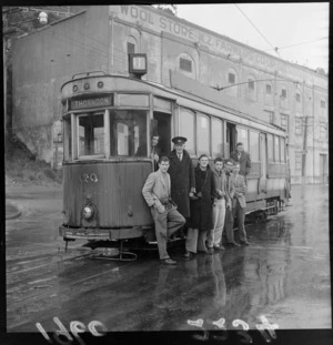 Tram driver with passengers standing outside tram hired for sightseeing, on Hutt Road, Thorndon with Wool Store New Zealand Farmers Co-op building behind, Wellington