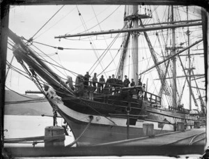 View of the prow of the sailing ship 'City of Lahore' berthed at Port Chalmers, showing her figurehead and group of crew members in the bow, looking down at the camera below, 1881.
