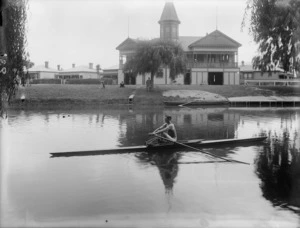 Single sculler on the Avon River in front of the Christchurch Amateur Rowing Club building, with men looking on, Christchurch