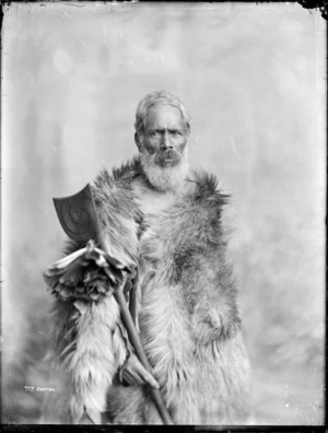 Unidentified Maori man wearing a cloak and holding a tewhatewha - Photograph taken by Frank Denton