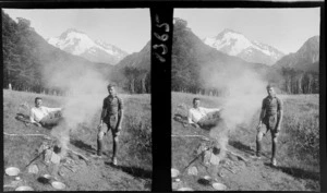 [Darran Mountains?], two unidentified men on grassed river flats beside a camp fire with forest and [Mount Madeline?] beyond, Fiordland National Park, Southland Region