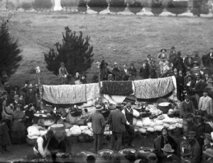 Crowd surrounding a rack of Maori cloaks and a pile of produce
