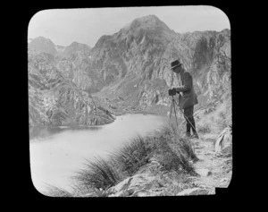 William Williams with a camera taking a photograph, beside small lake with mountains behind, unidentified location, South Island