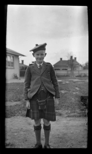An unidentified boy dressed in Scottish kilt, sporran, socks and hat in front of houses at an unknown location, [Dunedin City]