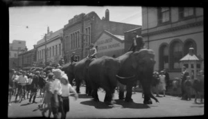A group of elephants ridden by unidentified men walking down a street in front of Turnbull & Jones Ltd Electrical Engineers building with a crowd of people looking on, [Christchurch City?]