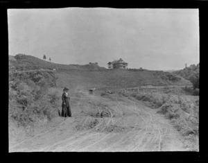 View of Lydia Williams on a dirt road in front of a hill with a two story wooden house surrounded by farmland, Kakahi District, Manawatu-Whanganui Region