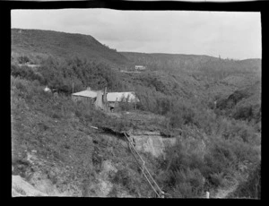 View of wooden houses and a road above a scrub filled gully with forest beyond, Kakahi District, Manawatu-Whanganui Region