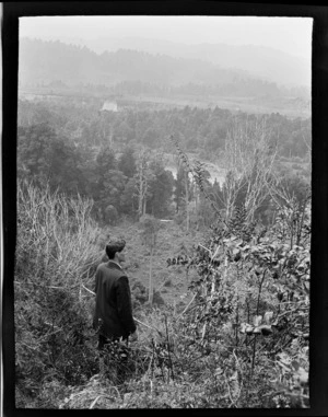 An unidentified young man on a scrub covered hillside looking out over a forest covered valley and the Whanganui River, Kakahi District, Manawatu- Whanganui Region