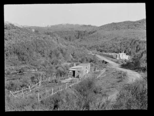 Cottages with fenced vegetable gardens and unidentified couple in doorway of one, next to unknown stream and scrub covered hills beyond, Kakahi District, Manawatu-Whanganui Region