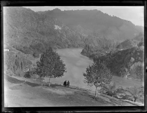 View over a lawn area of Pipiriki House with two unidentified people looking over the Whanganui River to riverbank houses and forest covered hills beyond, Pipiriki