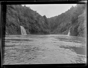 View off a boat on the Whanganui River with two side stream waterfalls down steep plant covered river banks with forest beyond