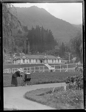 View of the Cadman Bathhouse (opened in 1898) on Te Aroha Domain with an unidentified woman, pushchair and child in front with a forest covered hill behind, Te Aroha, Waikato Region