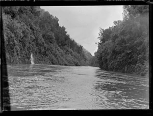 View off a boat on the Whanganui River showing waterfall down steep fern covered river banks and forest above