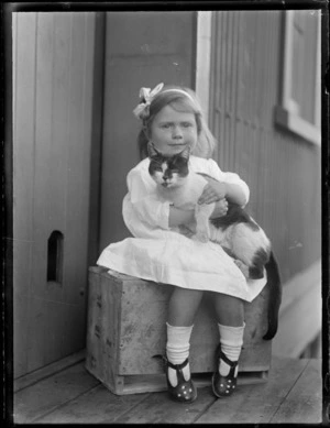 Portrait of an unidentified young girl sitting on a box outside an unknown house location holding a cat