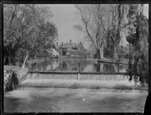 View of a weir across the Avon River with trees, a rowing boat and a large Tudor style house beyond, [Mona Vale], Christchurch City