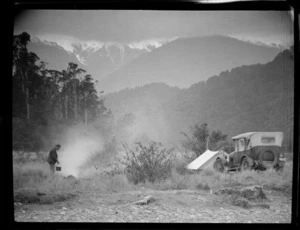 Unidentified man cooking at a campfire beside a car and tent, with beech forest and mountain range in the distance, Westland