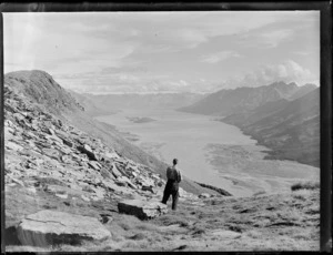 Edgar Williams looking down over the head of Lake Wakatipu, showing islands and mountains with schist rock to his left, Otago District