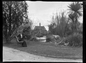 Unidentified woman sitting on park bench next to pond with the Bath House in the background, Government Gardens, Rotorua