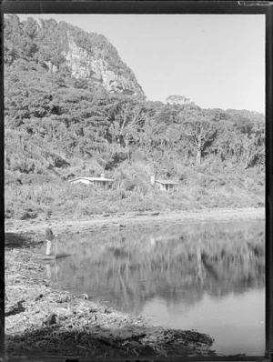 An unidentified man on an unknown beach with a rowing boat and two modern style batches behind, with forest beyond, Westport District, West Coast Region